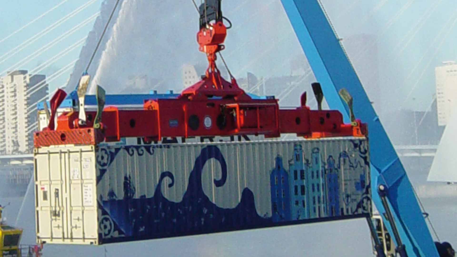 A large container hangs in the air on a container spreader with a city in the background