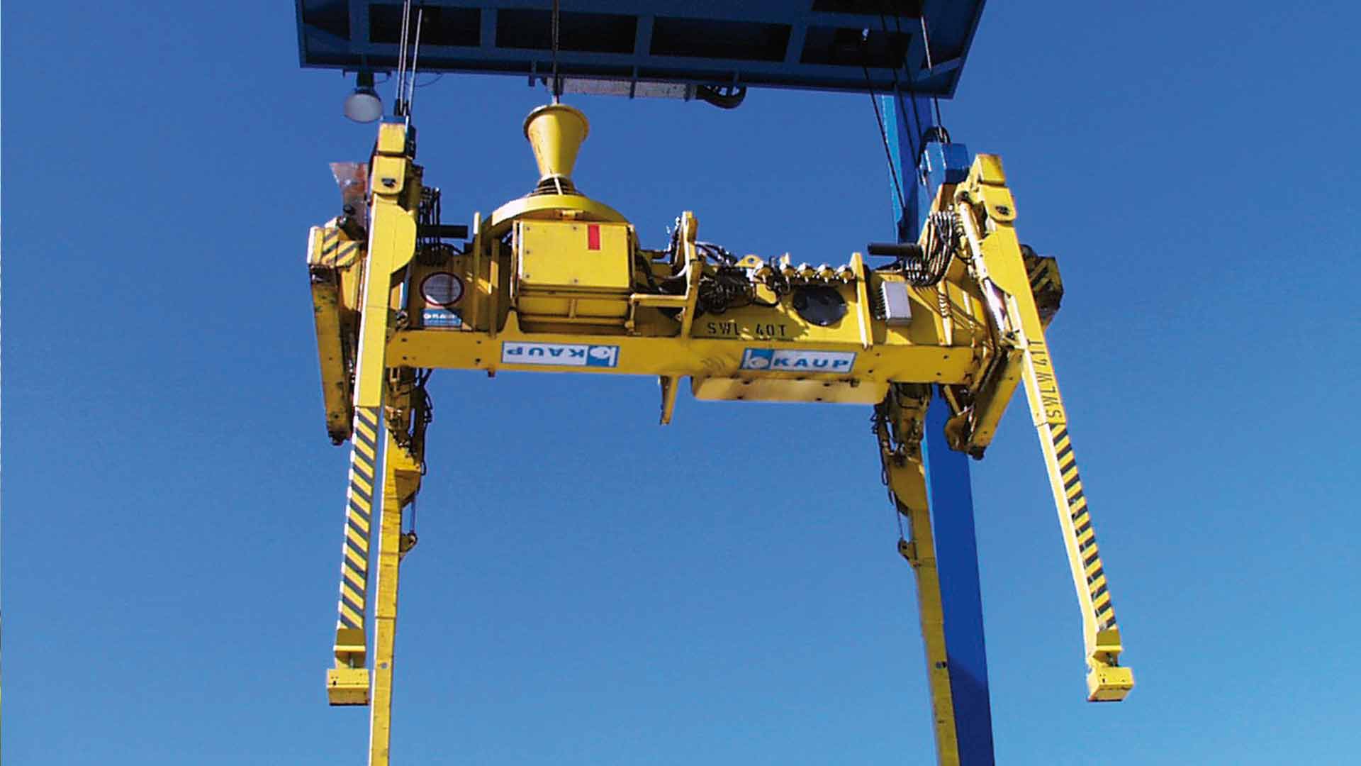 Large yellow container spreader from KAUP attached to a crane in front of a blue sky
