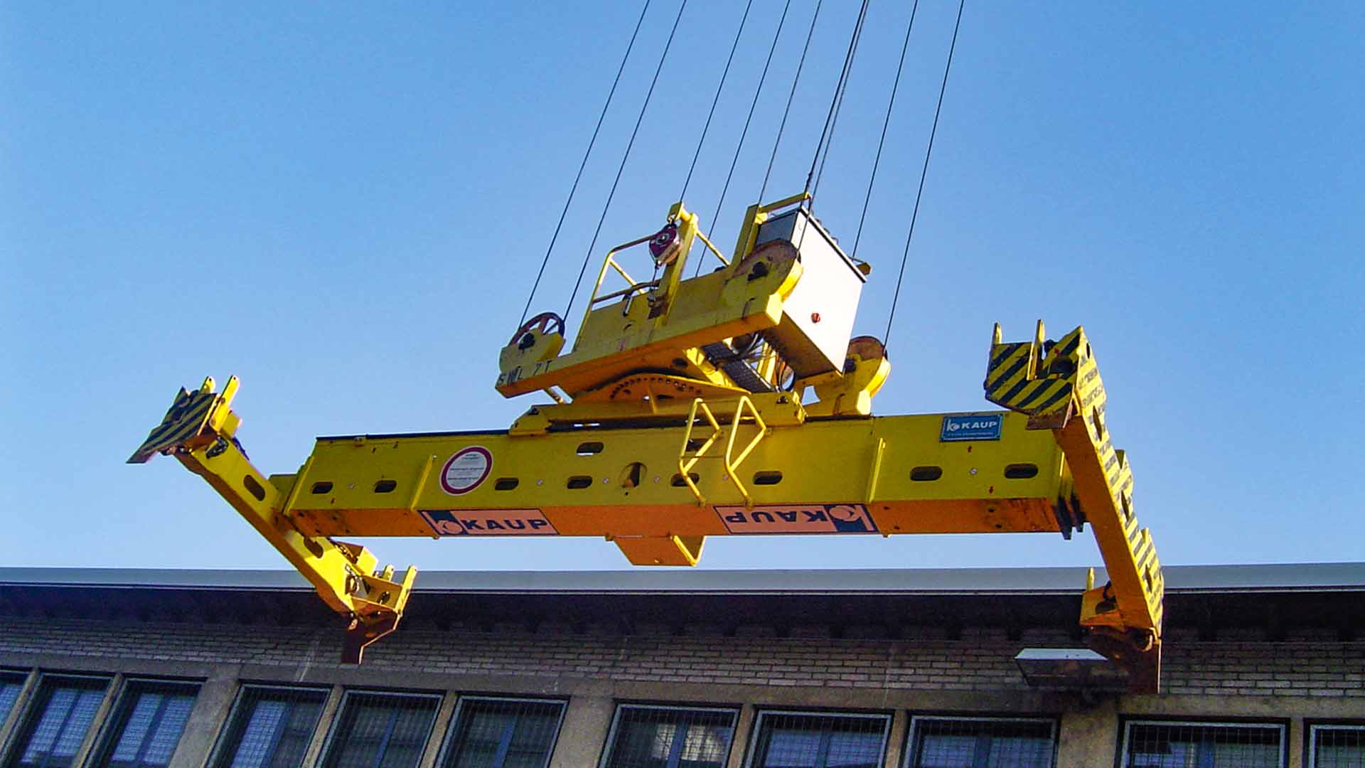 A yellow container spreader from KAUP hangs down from a crane on several wires