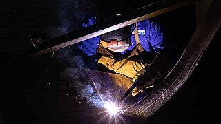 A welder in blue and yellow work clothes bends over a metal plate in a dark environment