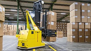 A yellow driverless transport system lifts up three stacked cartons in a warehouse