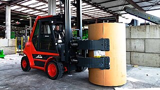A red forklift truck transports a large cylindrical cardboard roll with a roller clamp