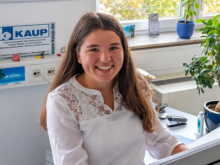 Eine junge Frau mit weißer Bluse sitzt im Büro an einem Schreibtisch und lächelt in die Kamera