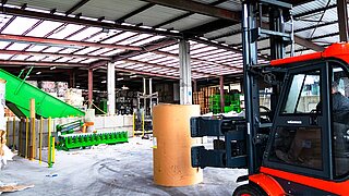 A forklift truck with a roll clamp drives towards an upright roll of paper in a warehouse