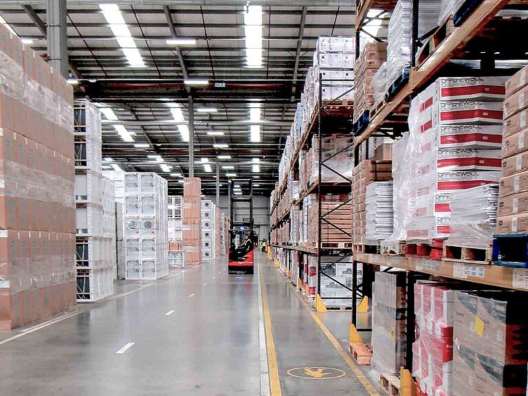 A forklift truck drives along an aisle with filled storage racks and stacks of boxes