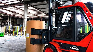 A red forklift picks up a cylindrical paper roll with a bale clamp for transport through a warehouse