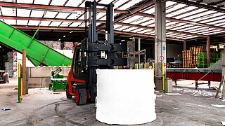 A red forklift truck with an open bale clamp drives towards a roll of paper standing on the ground