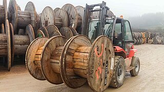A construction machine transports three large wooden rollers side by side with an attachment