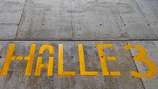 Grey concrete slab floor with the yellow, rounded, permanent lettering "Halle 3" on it