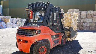 A red construction machine transports several stacked bales of waste paper with a mounted implement