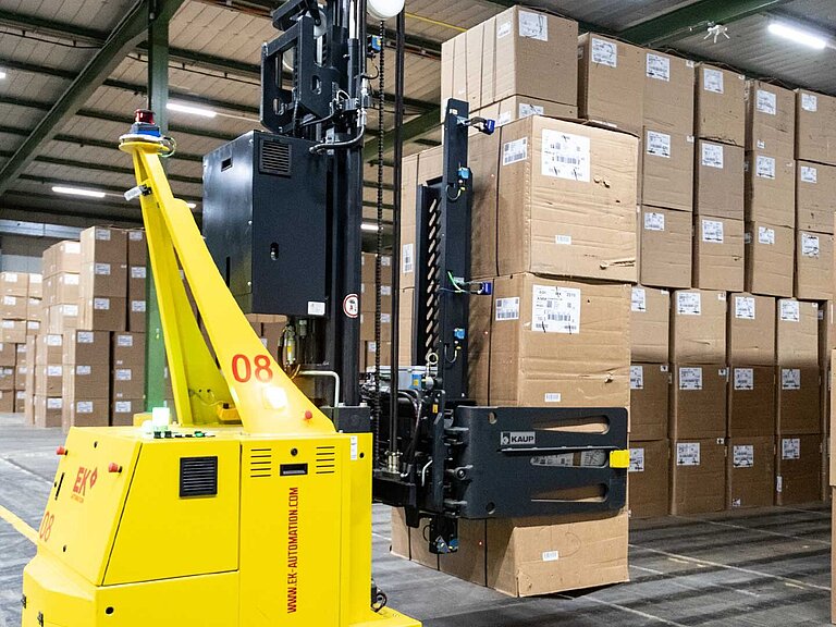 A yellow driverless transport system lifts up three stacked cartons in a warehouse