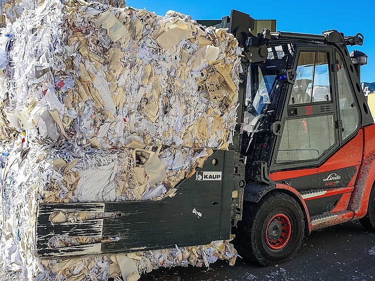 A red forklift truck transports two large pressed bales of waste paper with a clamp