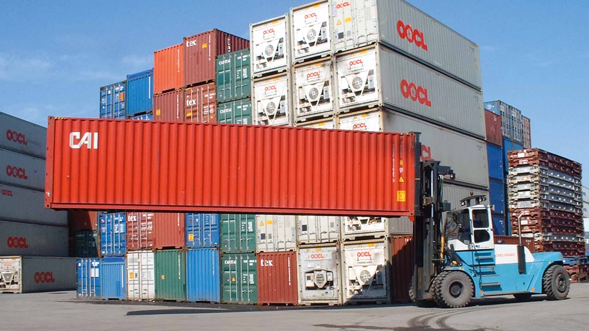 A forklift truck lifts a red container on its long side over a container storage area